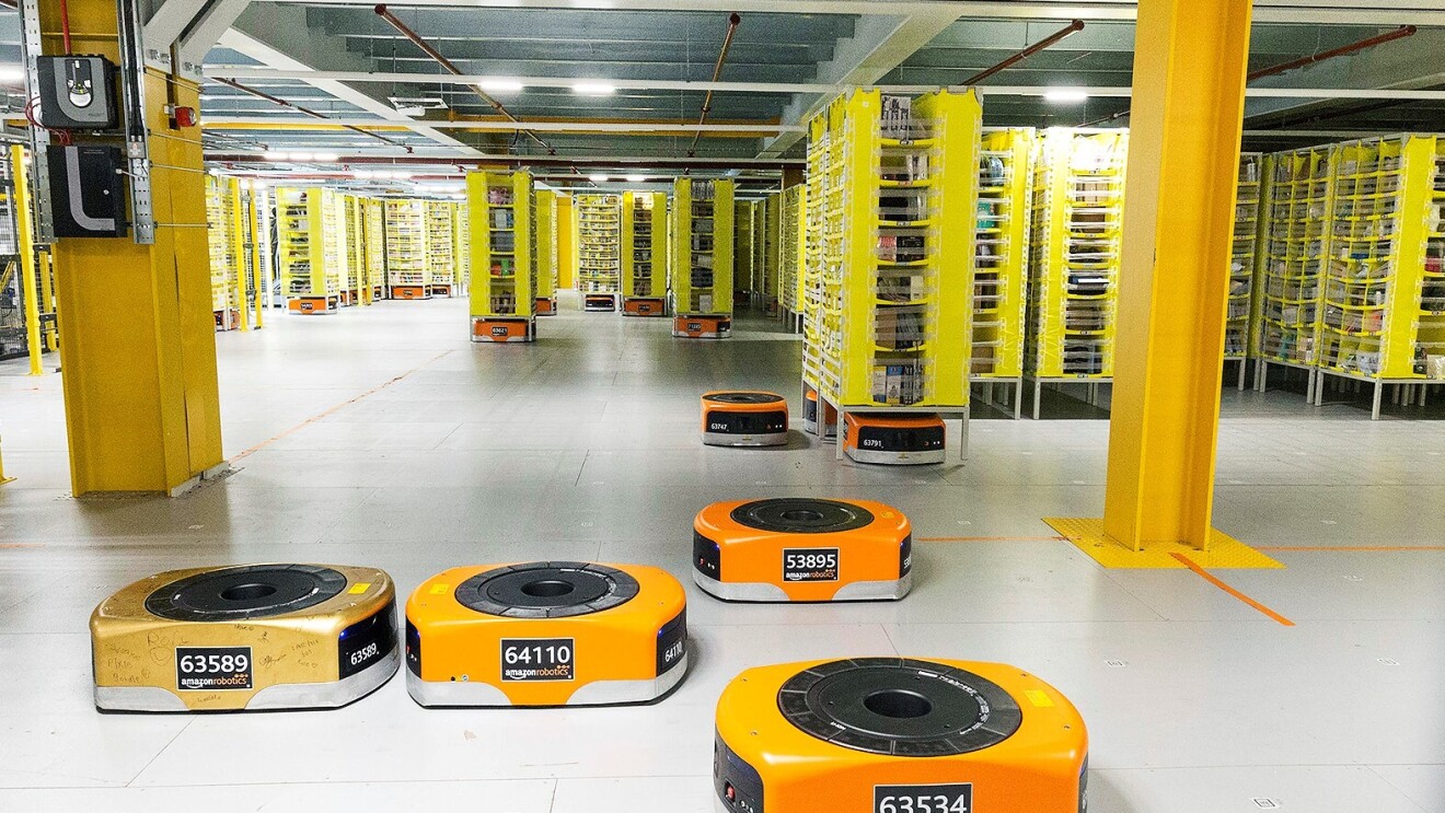 Several orange robots are pictured in a Fulfillment Center as they help move products across the floor.