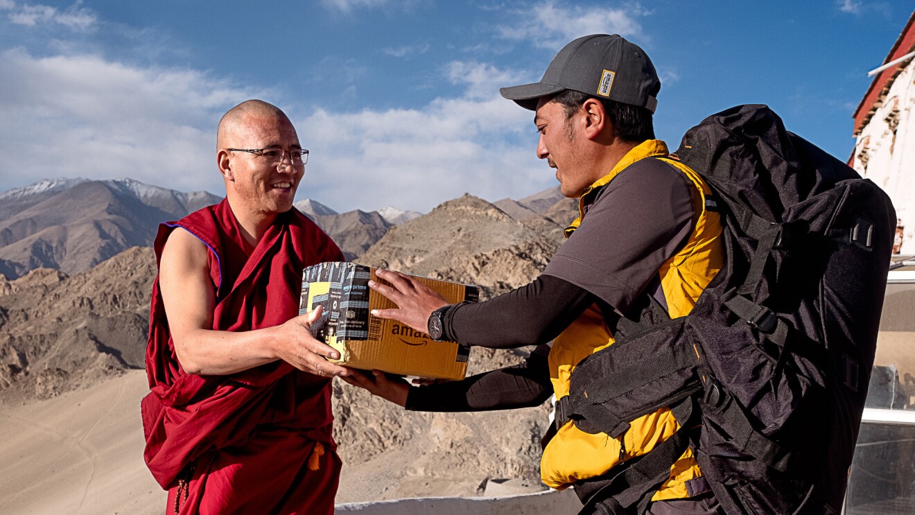 A delivery driver hands an Amazon package to a monk. Behind them, mountains and blue skies are visible.