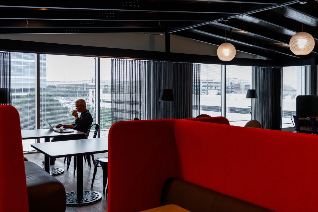 Inside the Amazon office in Austin, Texas, an Amazonian sits at a table near the window, next to him is an empty table. In the foreground are high-back red booths.