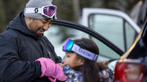 A father smiles as he looks down and helps his young daughter get her winter glove on.