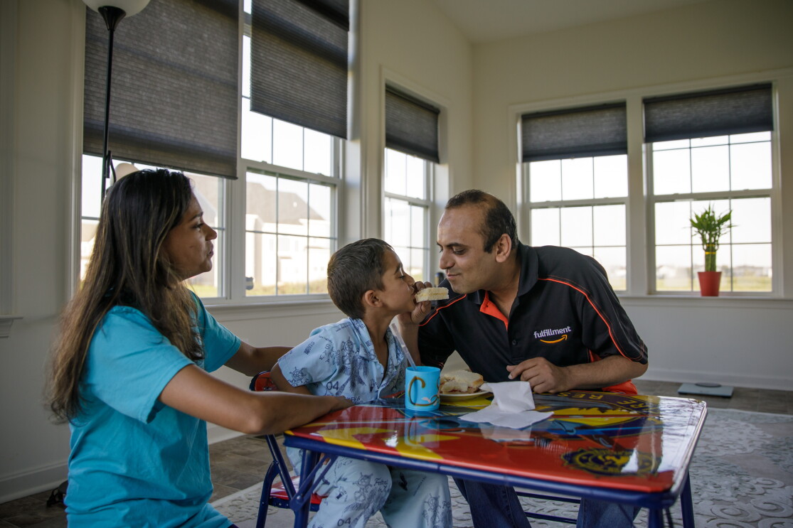 A woman, a young boy, and a man share a meal while sitting around a table in a room with several windows. The man holds food in his right hand and offers it to the boy, who takes a bite.