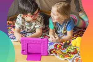 An image of two children laughing and interacting with an Amazon tablet. They are laying down on a colorful carpet and have toys sitting on the floor around them. 