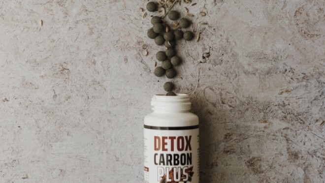 Una bottiglietta di integratori Detox Carbon Plus rovesciata su un piano. Dal collo della confezione escono ingredienti naturali.