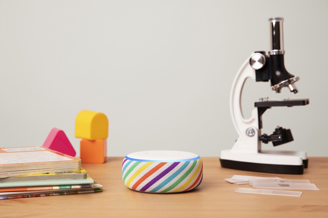 Amazon Echo Dot in rainbow stripe, displayed on a wooden tabletop. To the left are books and blocks, to the right is a microscope.