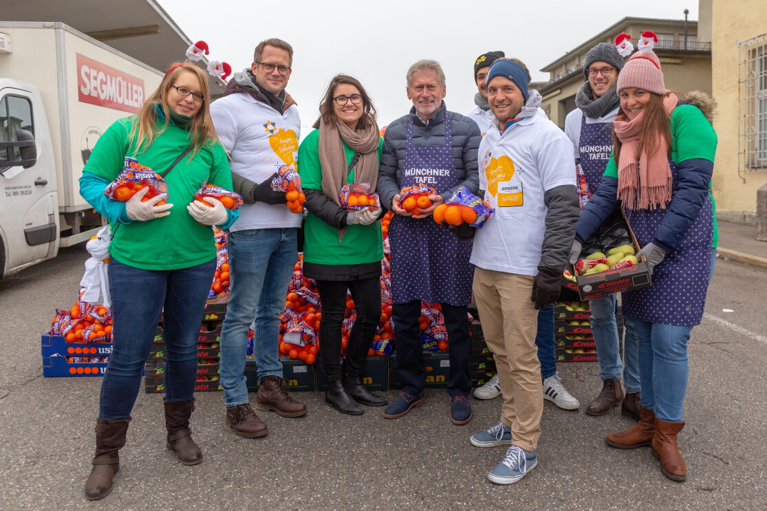 Eine Gruppe von Amazon Mitarbeitern, u. a. von Fresh mit grünen Shirts, der Rest mit weißen Shirts, steht vor einem Berg von Orangen und Mandarinnen. In ihrer Mitte: Paul Breitner.