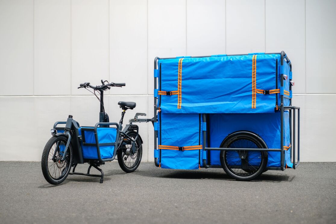 自転車にとり付けられた青いバスケットと青いカバーが付いた大きなリアカーが白い壁の前に置かれている。リヤカーの荷台のカバーには落下防止用のオレンジ色のベルトが取り付けられている。