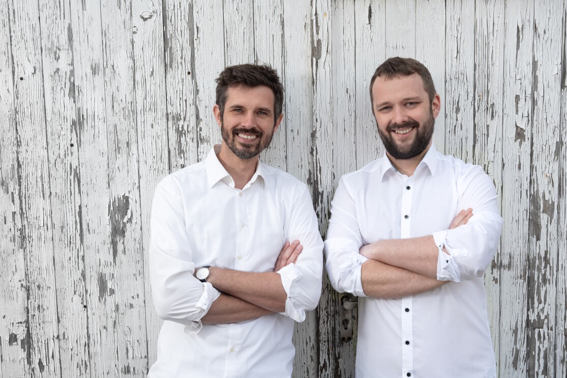 Zwei Männer mit dunklen Haaren und weißen Hemden stehen vor einer Wand und lachen in die Kamera.
