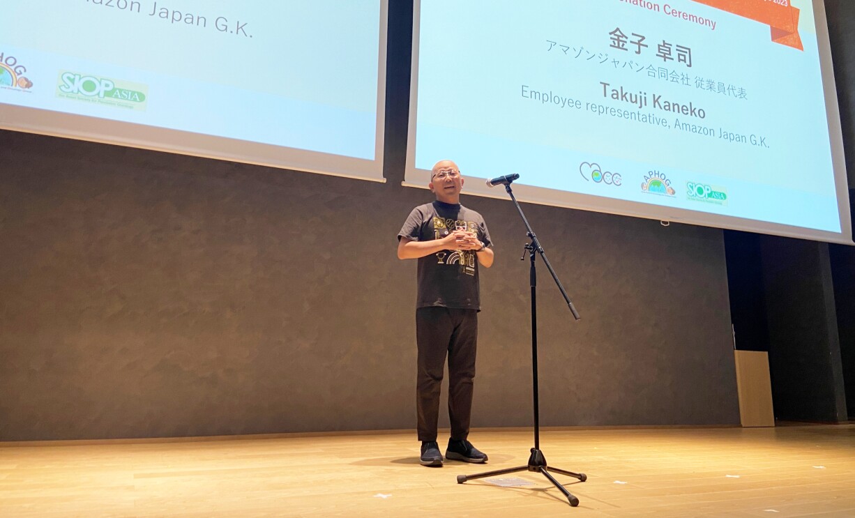 ステージに立ちマイクの前で話しているメガネをした男性