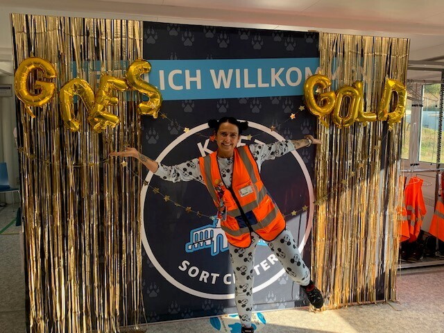 Eine Mitarbeiterin steht im Schlafanzug im Sortierzentrum in Wolfhagen vor einem Bild auf dem "Amazon goes gold" steht.