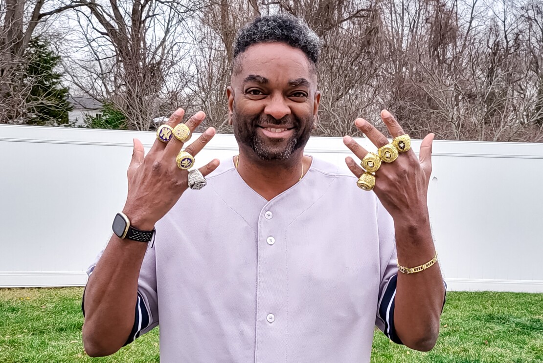 An image of a man smiling for a photo in his backyard. He is holding up both hands to show the championship rings on each of his fingers. 