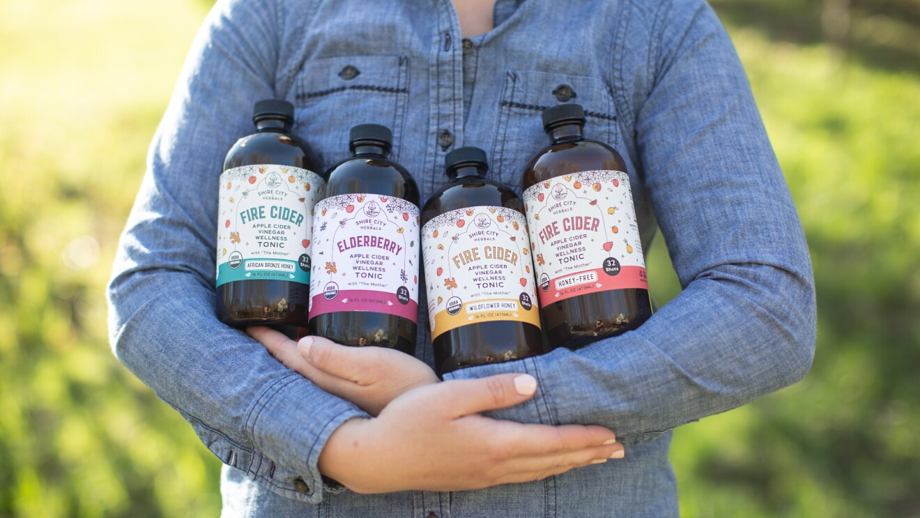 A person holds four bottles of Fire Cider in their arms while standing outside.
