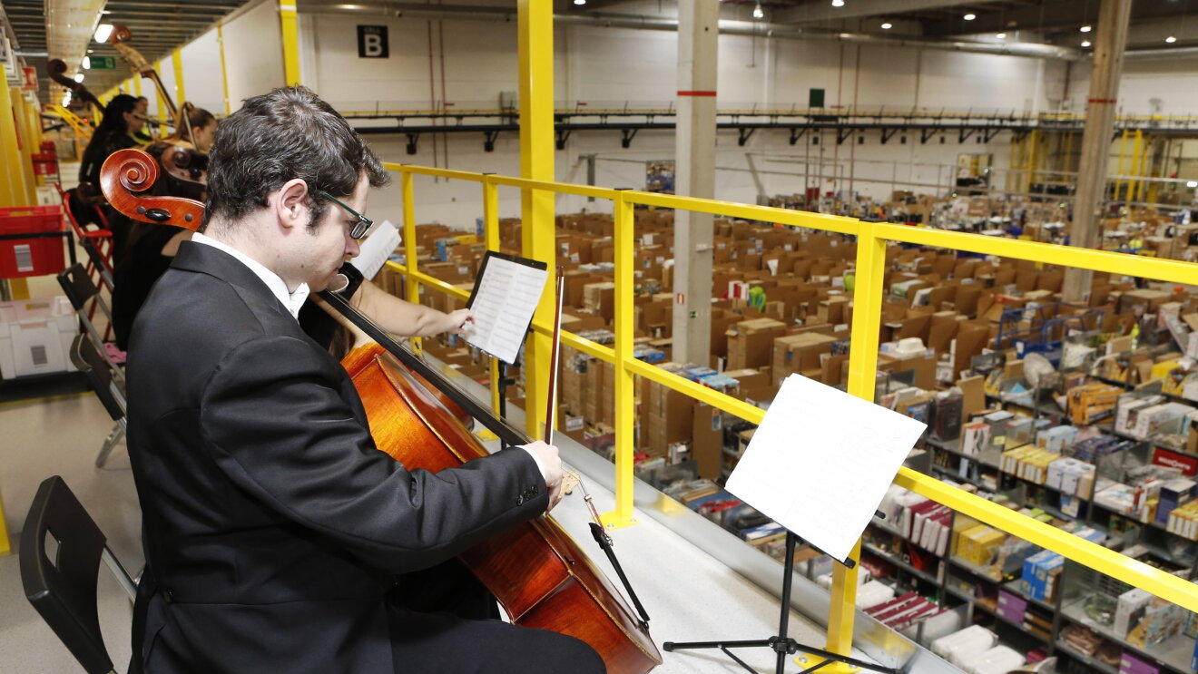 Hombre de mediana edad, con gafas, tocando el violonchelo con la mano derecha. La acción se desarrolla en un centro logístico y detrás de él se adivinan más instrumentos de cuerda. Delante de él se observa, a un nivel inferior, una planta llena de productos.