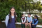 A young girl smiles and poses for the camera while three of her classmates sit behind her smiling at one another. They are outside surrounded by greenery and wear school uniforms.