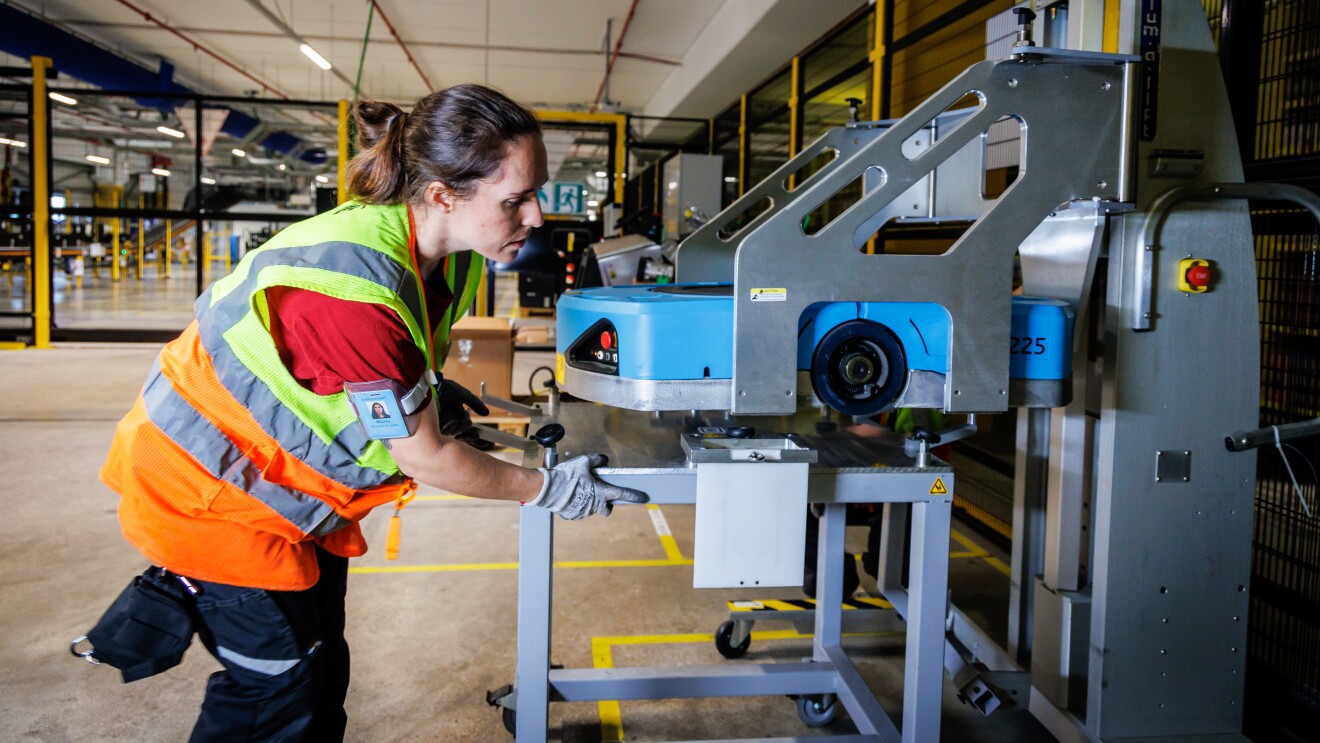 Dentro de un centro logístico, está una mujer con un elevador para reparar un robot de color azul. Está de pie, lleva el pelo recogido con un moñparrodillada mirando un robot está colocado en una mesa. Ella tiene el pelo oscuro largo y recogido en una cola. Van con un camiseta negra de manga corta y un chaleco amarillo.