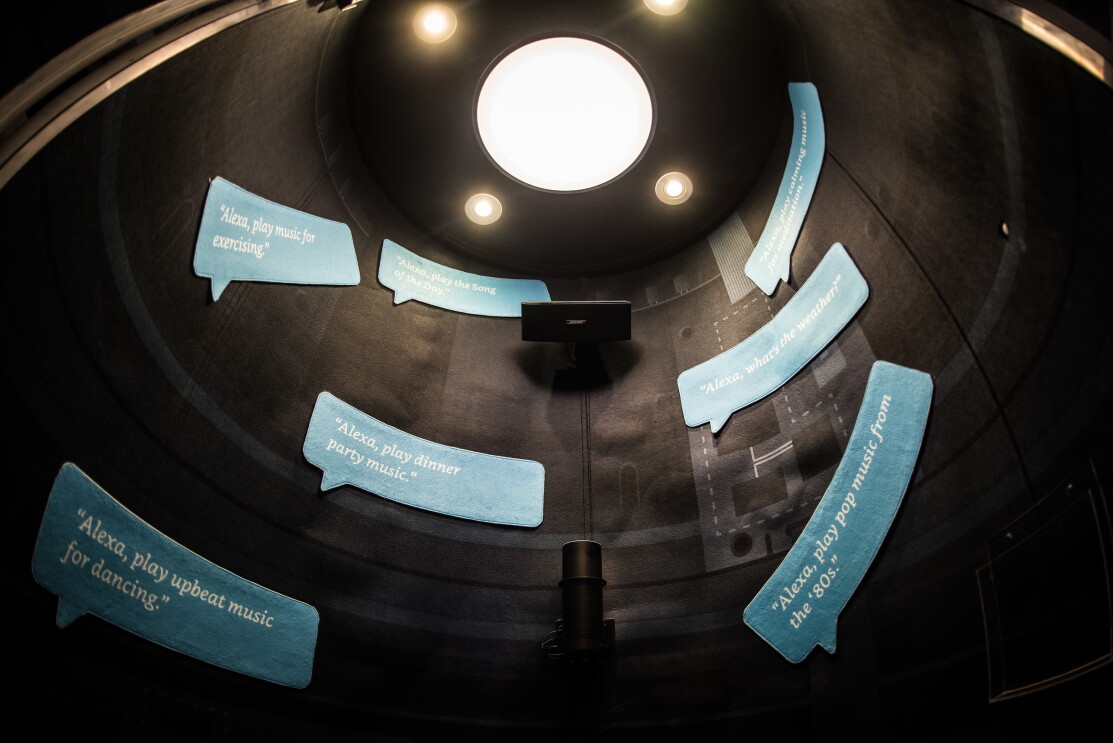 Día Europeo de la Innovación 2018. Construcción cilíndirca de color negro con cuatro focos que iluminan mensajes en ingles en bocadillos de cómic que hablan sobre Alexa. Uno de los mensajes es así: Alexa interpreta música de los '80, etc. 