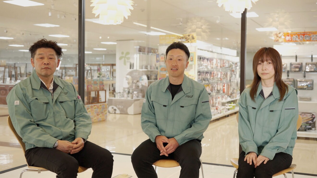調理商品の明るいショールームに、3名のスタッフが並んで座っている。ブルーグレーの作業服を着用し、中央の人物は手を軽く組んで話をしている。背景には商品棚や照明が見える。