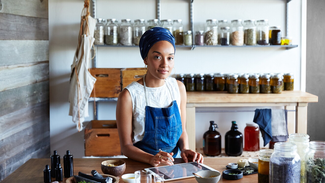 A woman stands at a table, working on an iPad. Behind her are jars of preserves or other homemade products, and there are herbs and ingredients in front of her.
