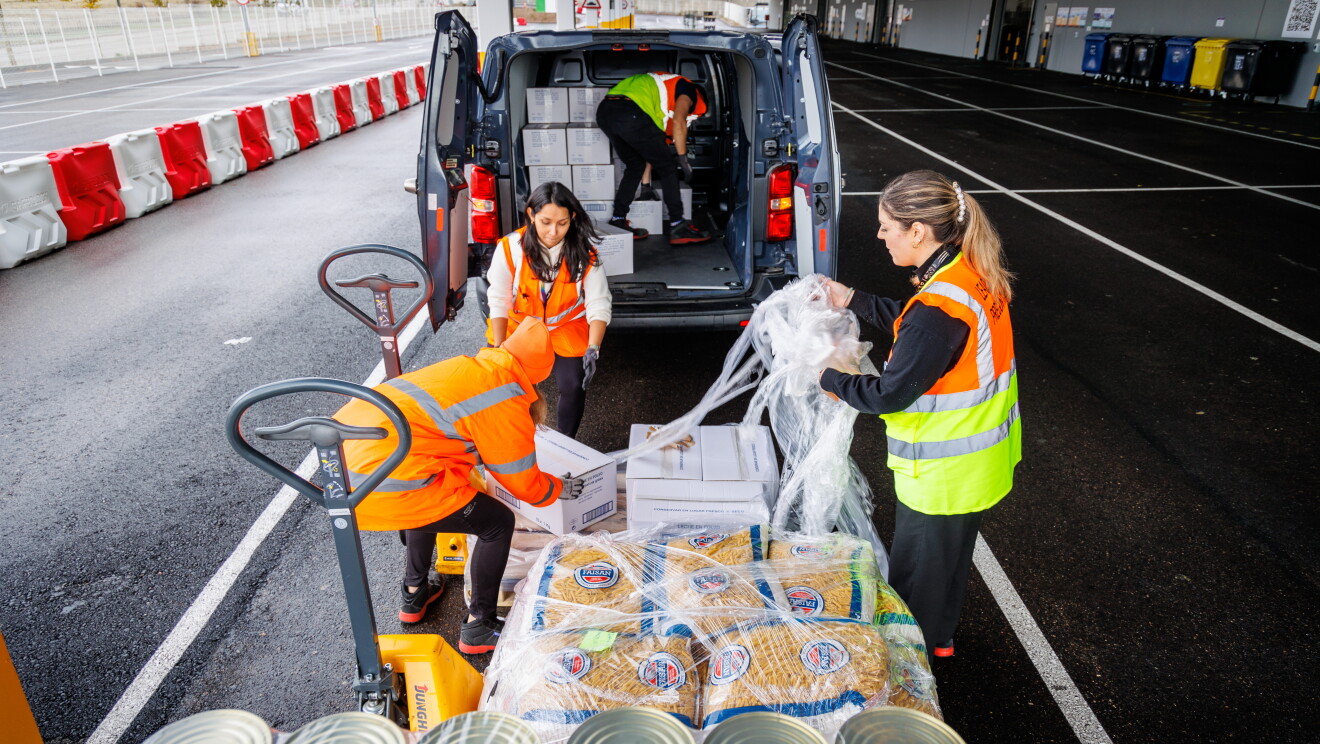 En primer plano latas de comida y paquetes de macarrones. En el margen izquierdo se ven dos toros semiautomáticos. Tres mujeres quitando el plástico transparente que envuelve los palés. Dos de ellas van con chaleco naranja y jersey debajo y la tercera lleva un abrigo de color naranja. En el fondo se ve una furgoneta con el maletero abierto y semilleno. El hombre va con chaleco, guantes y manga corta.