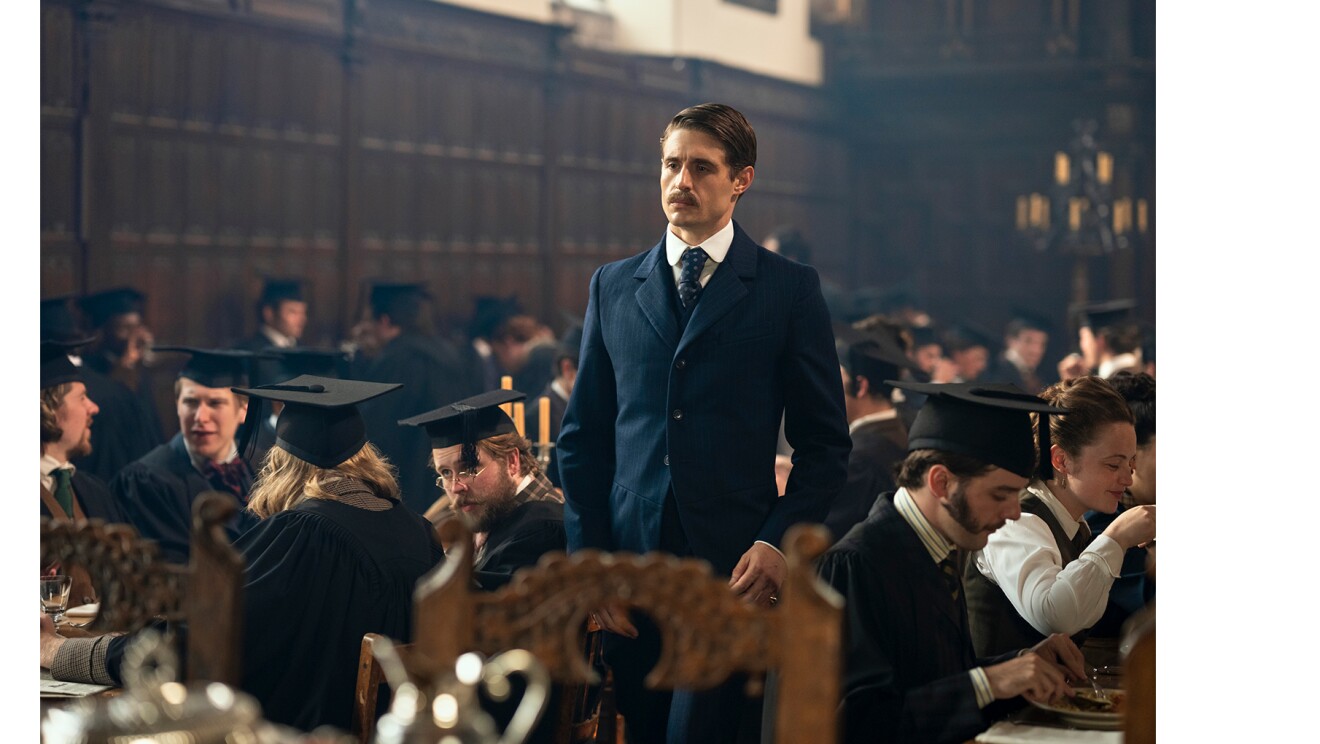 四角い学帽と黒いガウンを着用した古風な学生たちが大勢テーブルについて食事をしている食堂。口髭を生やしたスーツの男性がひとりだけ立っている。