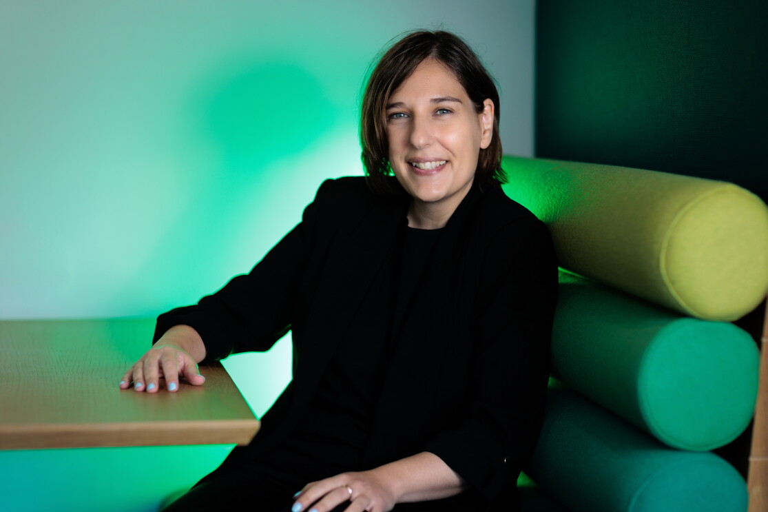 María Alonso vestida con un traje pantalón de color negro, con un jersey negro también debajo de la americana. Con el pelo corto y los ojos azules. Las manos con las uñas azules. Con la mano derecha apoyada en la mesa y la otra en la pierna. Sentada en un sofá con tonos verdes y luces verdes de fondo. 