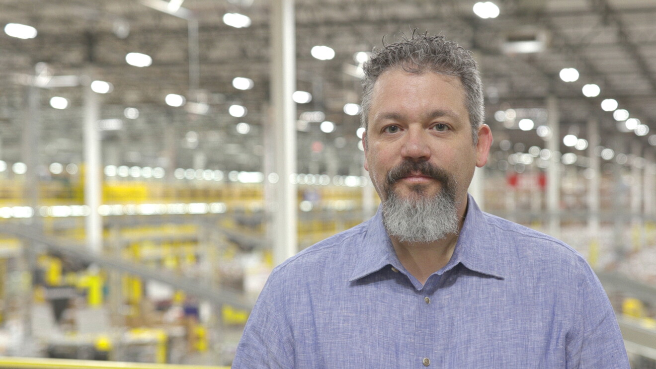 Portrait of a man in a light blue button down shirt looking at the camera. The man's hair and goatee are a mix of white, gray, and black. A large warehouse space can be seen in the background.