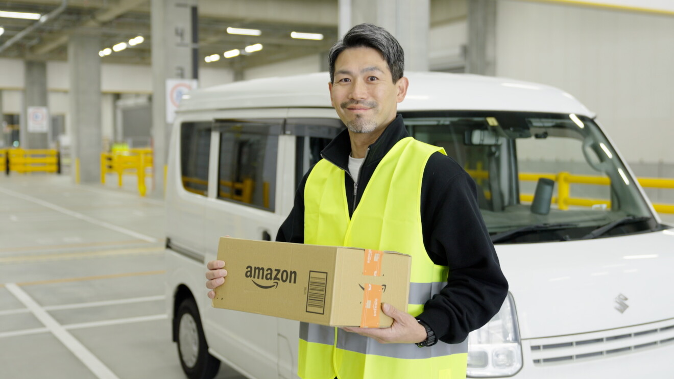 駐車場に駐車している白い軽自動車の前に立ち、段ボールを手に持つ男性