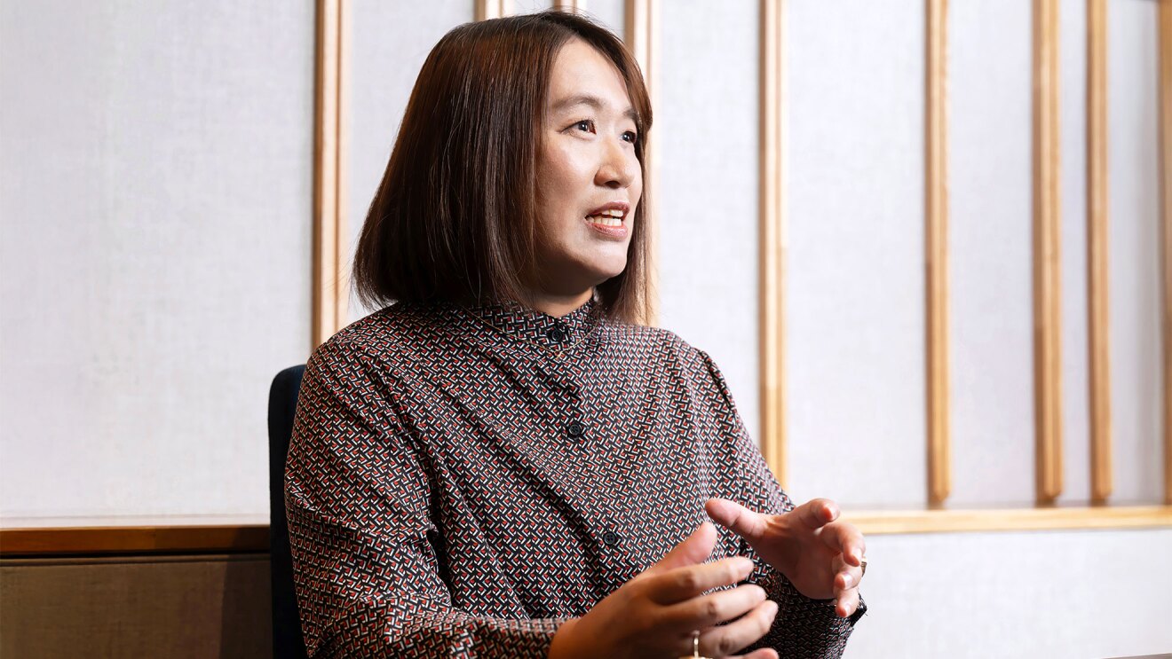 ベージュの壁に、木製の仕切りが複数配置された明るい室内で、椅子に座り、赤と紺の細かい幾何学模様のワンピースを着た女性が体の前に両手で形を作りながら話している