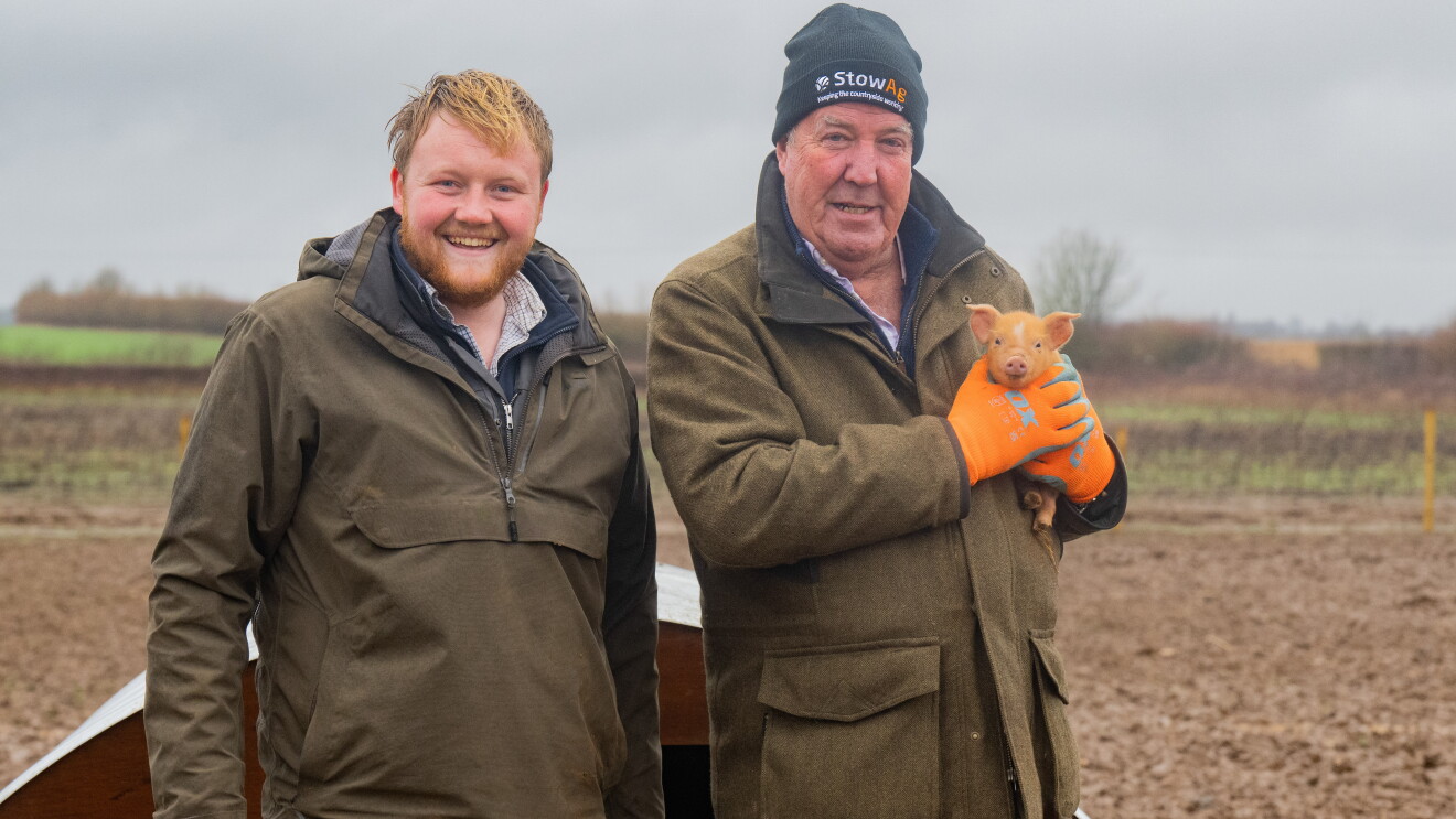 Two men holding a small pig with a farm in the foreground 
