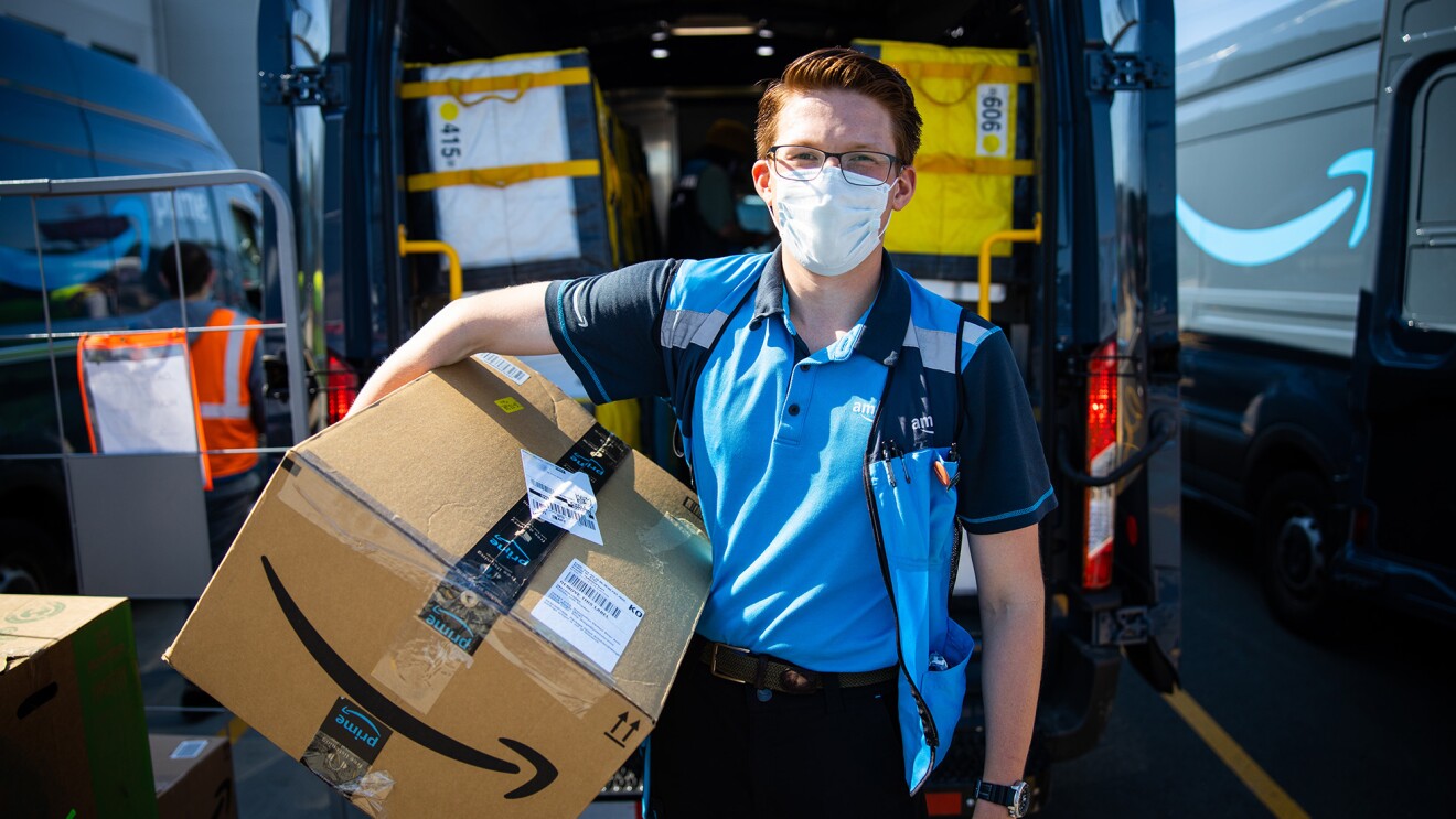 An Amazon delivery driver stands for a photo in front of his delivery van, holding a box and smiling under a mask. 