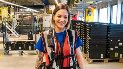 Alba Muñoz, asociada en el área de robótica, con sus herramientas de trabajo. Sonriendo mirando a cámara, viste con su chaleco de trabajo lleno de bolsas llenas de utensilios. También viste con una camiseta azul y un pantalón tejano. De fondo está un pasillo del centro logístico del Prat de Llobregat. Castaña y con media melena tiene un piercing en la nariz. 