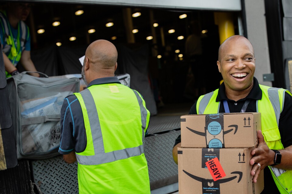 A smiling man holds boxes while people in the background load a truck. 