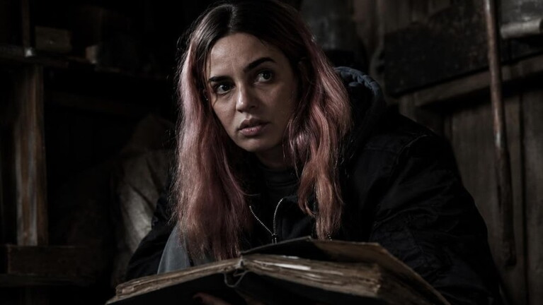 Woman with pink hair holds a tattered book in a darkened room, looking out with a scared expression 