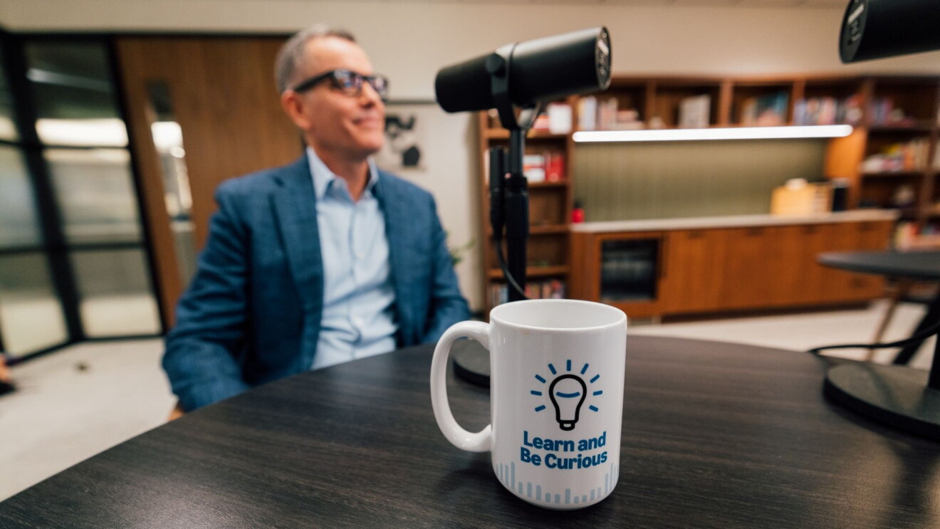 Podcast interview scene featuring Amazon Stores CEO Doug Herrington wearing a blue suit seated at a table with a prominent 'Learn and Be Curious' coffee cup.