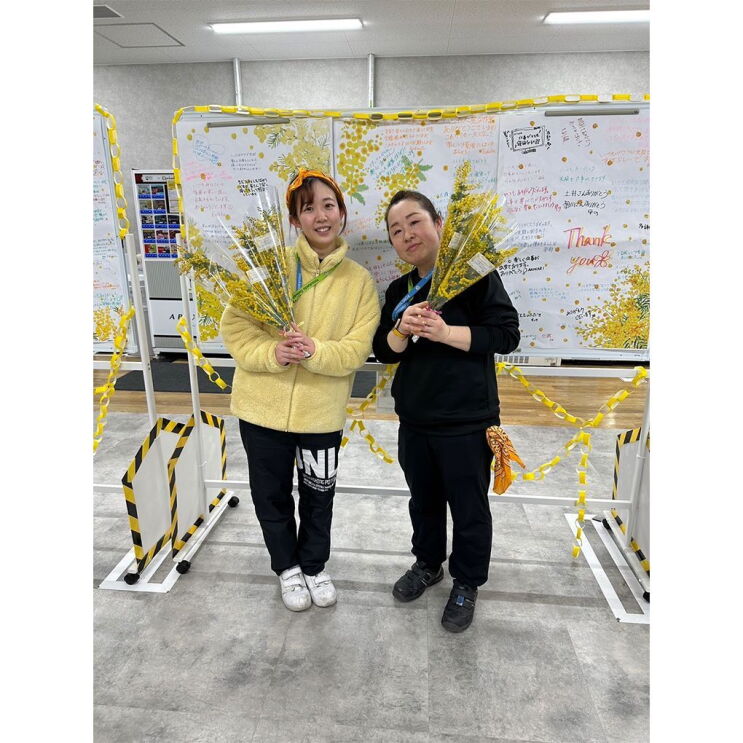 2人の女性がメッセージが書かれたホワイトボードの前に立って、黄色い花を持っている。
