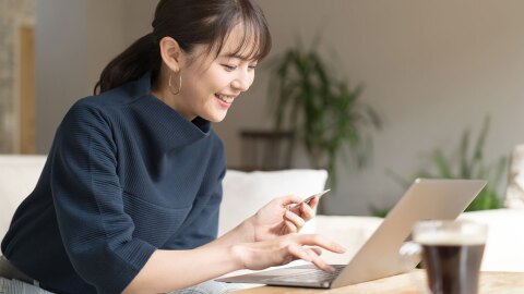 紺色トップスを着た人物が白いソファに座り、ノートパソコンの画面を見ながら微笑んでいる。左手にカードを持って右手でキーボードに触れている。明るい室内で、背景に観葉植物が見える。手前に黒い飲み物の入ったグラス。