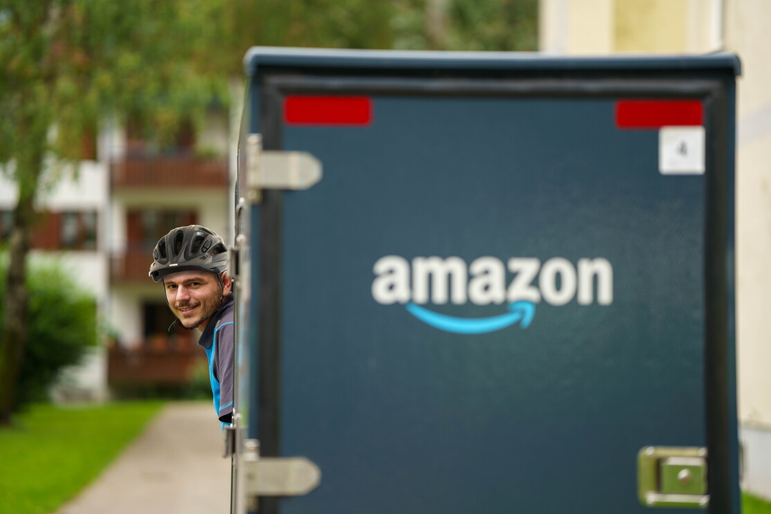 Ein junger E-Cargo Fahrer mit Fahrradhelm in seinem Fahrzeug, man sieht auf der Rückseite ein großes Amazon-Logo: Er lehnt sich seitlich heraus und blickt zurück, sodass man sein Gesicht erkennen kann.