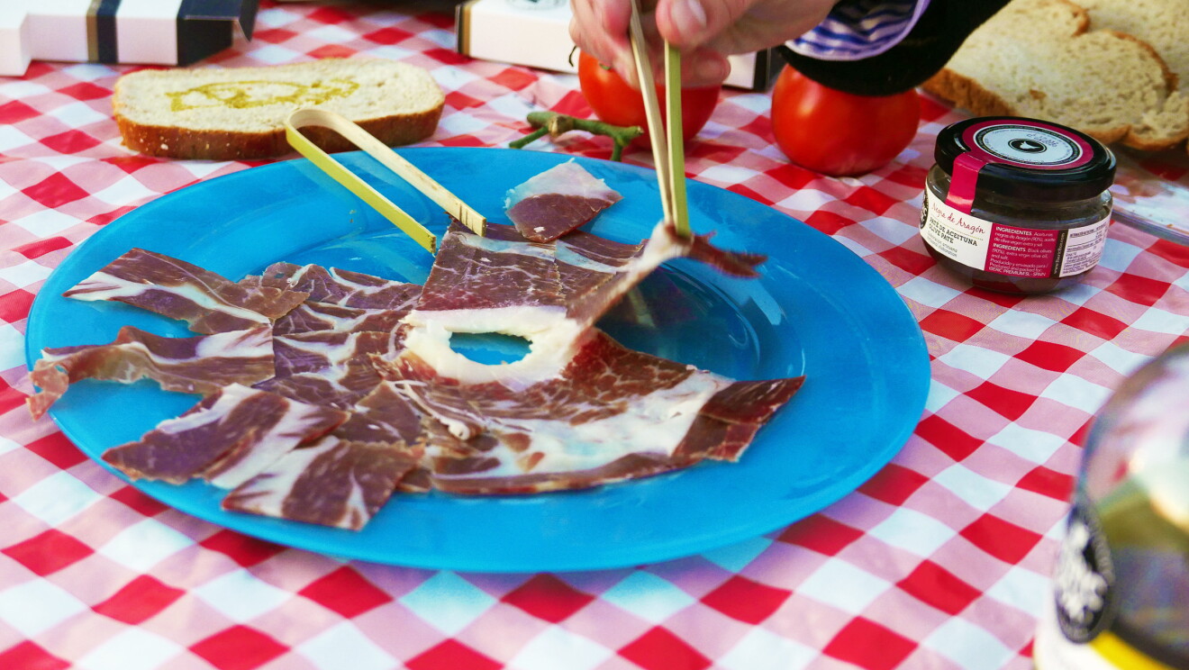 Primer plano de un plato jamón. El plato es azul y está sobre un mantel de cuadros rojos y blancos. Una mano con unas pinzas de bambú está cogiendo un corte. 
