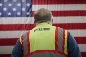 An image of an Amazon employee in a yellow work vest standing in front of an American flag.