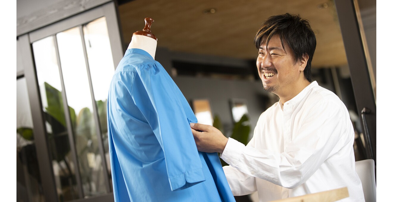 商品のブルーのシャツを手に笑顔の添田さんの写真