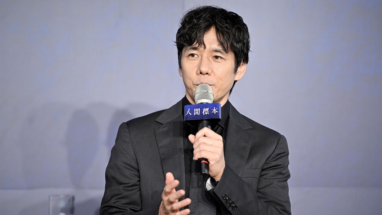 青みがかったグレーの壁を背景に、スーツ姿の人物の男性(西島秀俊さん)。スーツ、シャツ、ネクタイすべて黒を着用している。左手に持ったマイクには「人間標本」と書かれたパーツが付いている。