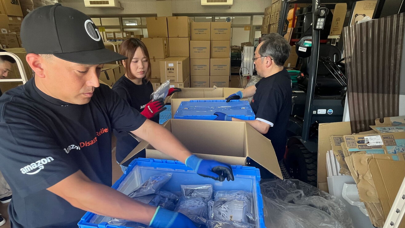 段ボールや資材が多く積まれた作業場で、荷物の箱を開けて作業をしている3人の人物。手袋を着用している。