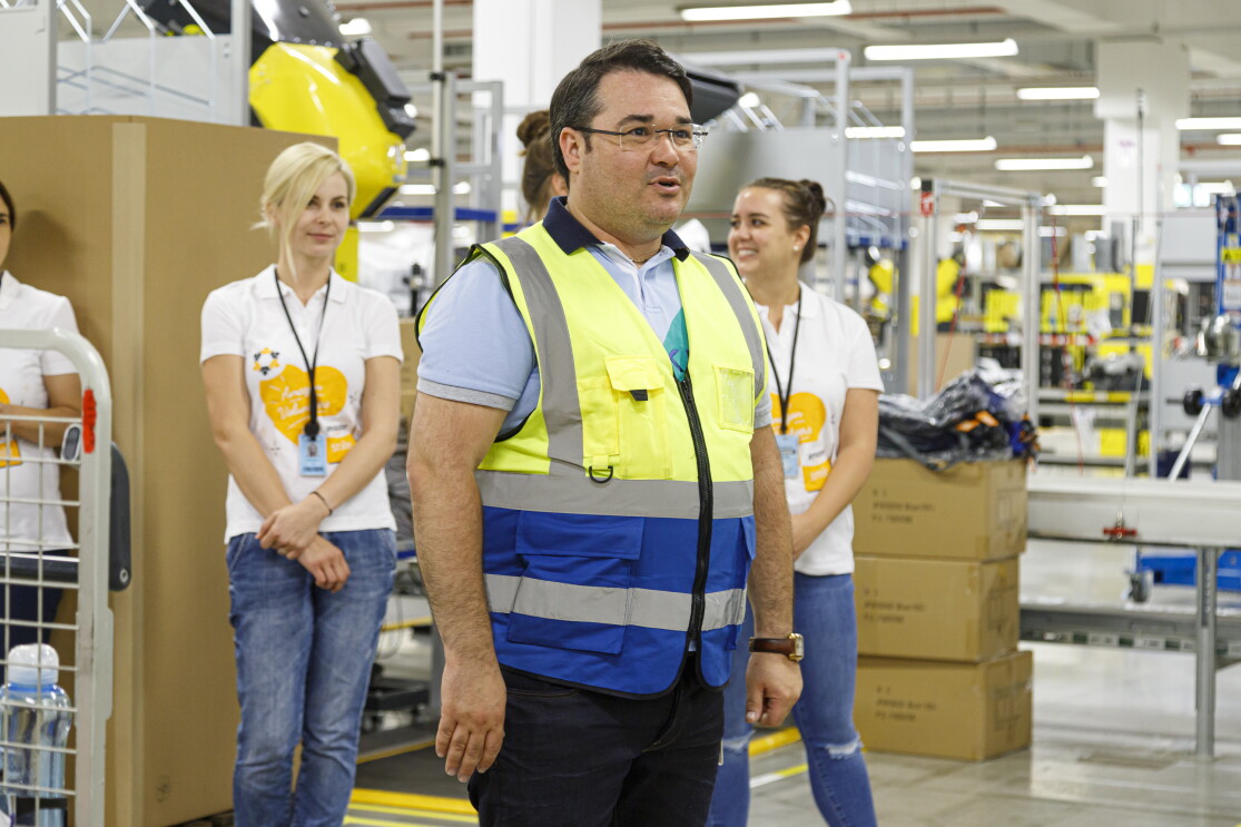 Ein Mann mit Brille, Sicherheitsweste und dunkelbraunen Haaren in einer Logistikhalle. Im Hintergrund sieht man 2 Mitarbeiterinnen von Amazon in "Amazon gemeinsam"-Shirts.