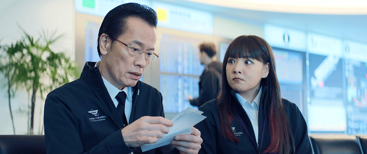 ロビーのような室内で黒いソファに並んで座る、中年男性と若い女性。2人とも濃紺の制服とシャツを着用。男性は数枚の紙を持っている。