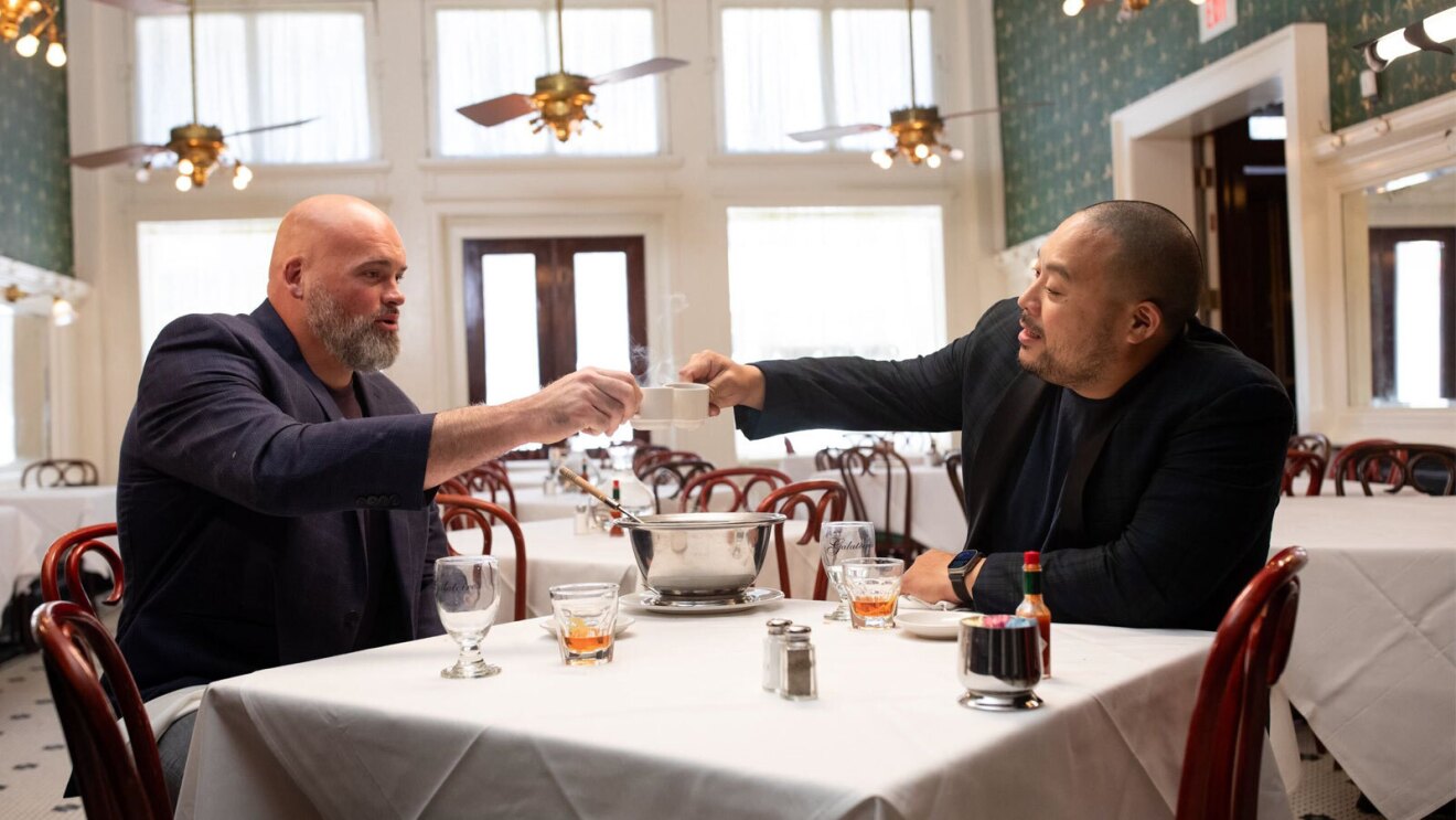 An image of two men sitting at a restaurant table. They are holding up espresso cups to cheers one another. 