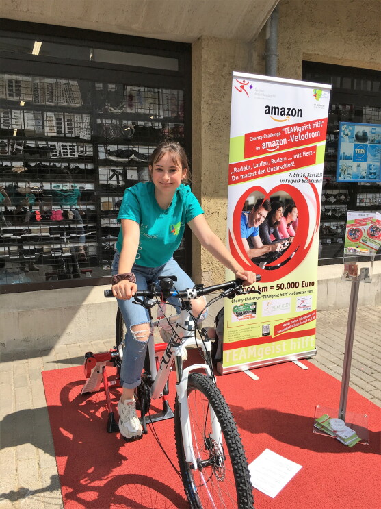 Eine junge Frau sitzt auf einem Trimmrad. Im Hintergrund steht ein Roll Up, das auf die Charity Challenge und Amazon weist.