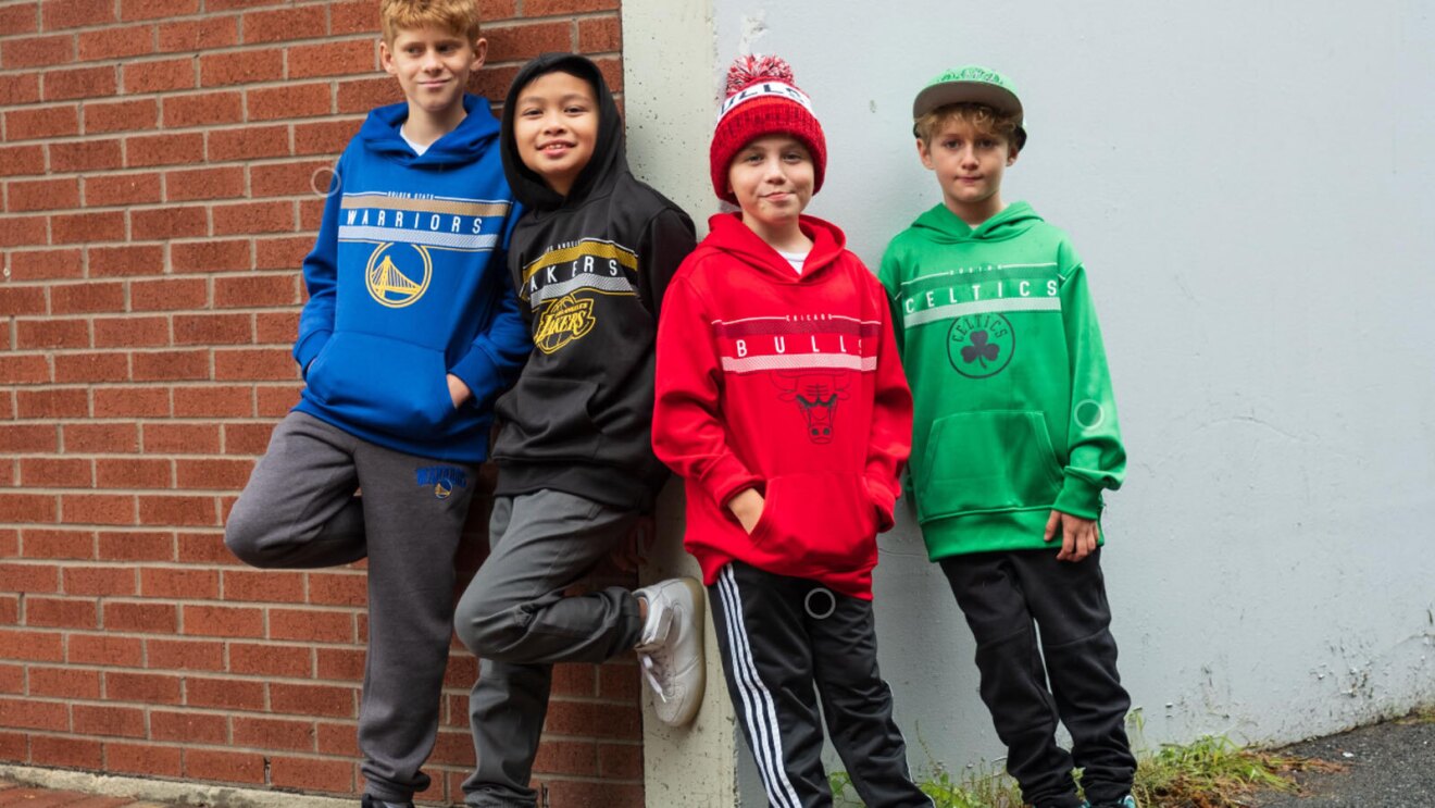 An image of four kids leaning against a brick wall and smiling for the camera. They are all wearing different NBA hoodies; A blue Warriors hoodie, a black Lakers hoodie, a red Bulls hoodie, and a green Celtics hoodie. Two kids have on hats; one red and black bulls beenie and a greek, flat-brim Celtics hat.