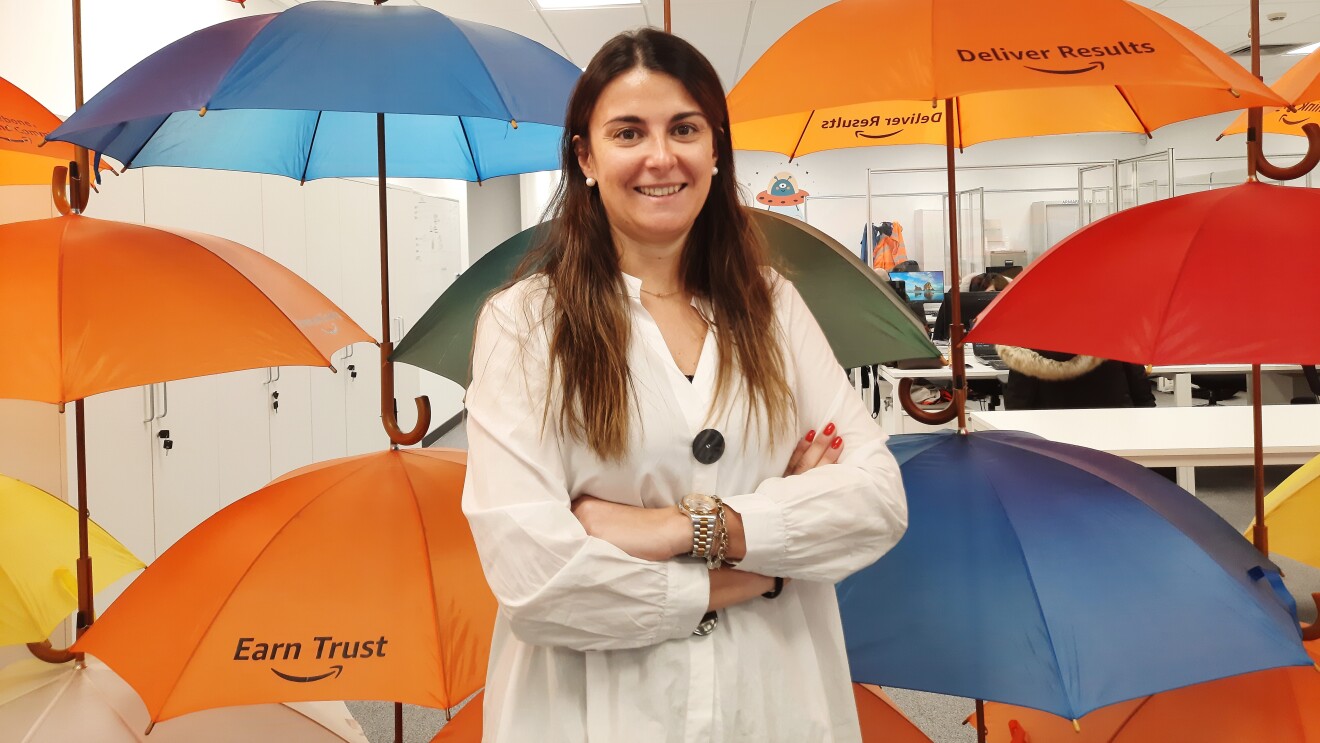 Raquel Mur. Va con una camisa blanca. Los brazos cruzados y mirando a cámara. Lleva las uñas rojas, unas pulseras en la mano izquierda. Tiene el pelo largo, liso y castañol. Mira a cámara sonriendo. El fondo es una oficina con paraguas que cuelgan del techo.