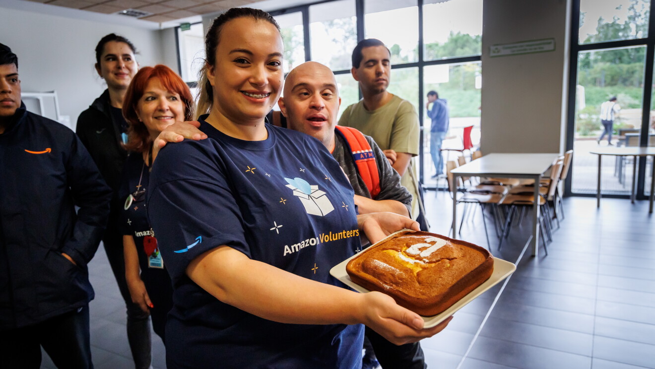 En las instalaciones de Down Madrid en Madrid aparece una trabajadora de Amazon con un bizcocho que tiene la letra A de Amazon y la sonrisa dibujadas con azúcar blanco. Detrás de la trabajadora un participante de Down Madrid agarrándola del brazo, De fondo lo que sería una cantina con meses y sillas.