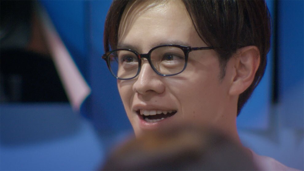 青い背景に、暗めの茶色のフレームの眼鏡をかけた人物のクローズアップ。口を開けた微妙な表情。手前には別の人物の頭部が一部写っているが、ぼやけており焦点が合っていない。