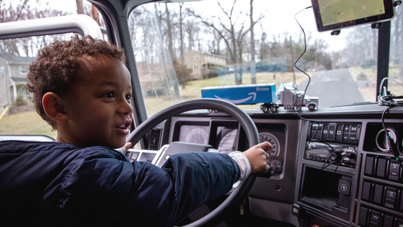 一个小男孩坐在一辆半挂式卡车的驾驶室里，双手放在方向盘上。他的玩具卡车就在方向盘前面。
